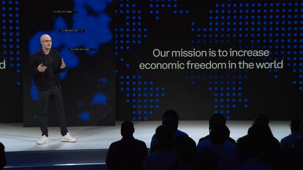 The image shows a man giving a speech or presentation, with a large screen behind him displaying text related to economic freedom in the world. The speaker appears to be addressing an audience of people sitting in front of them. The content displayed on the screen includes phrases such as "Our mission is to increase economic freedom in the world" and "It looks like @coinbase is trying to win on all fronts". These statements suggest that the man speaking might be discussing the impact of cryptocurrency or blockchain technology on global economies, with a focus on increasing economic freedom for individuals.