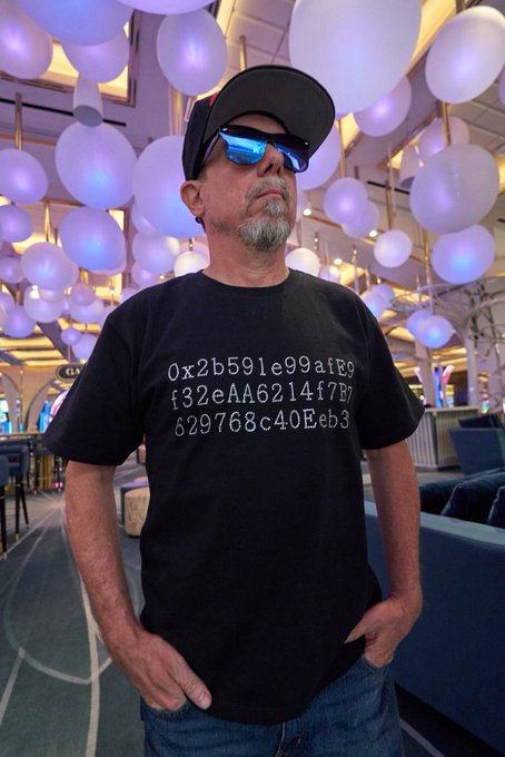 A man wearing a black cap, blue sunglasses, and a black t-shirt with a hexadecimal string printed on it, standing in a room filled with glowing spherical lights.