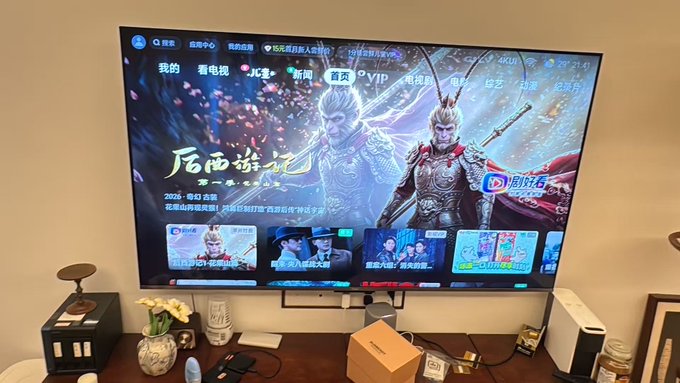 A wall-mounted flat-screen TV displaying a Chinese video streaming interface featuring a fantasy warrior show, with a wooden cabinet and various items below.