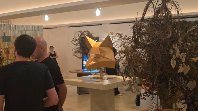 People viewing artwork at a gallery, including a yellow geometric fox-head sculpture on a pedestal and large dried floral installations in the background.