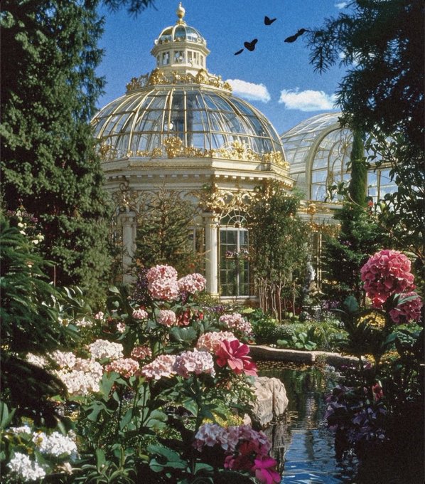 An ornate glass-domed conservatory surrounded by vibrant pink hydrangeas and a reflective pond.