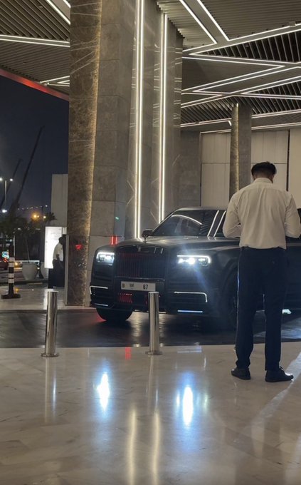 A black Rolls-Royce with illuminated headlights parked at a luxury hotel entrance, where a valet in a white shirt and black pants stands beside the vehicle.