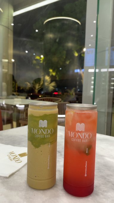 Two tall cans branded with 'MONDO COFFEE BAR' sit on a marble counter, containing green and red iced beverages.