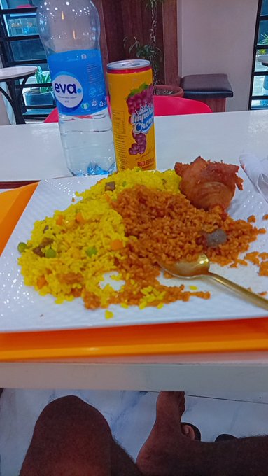 A plate of yellow and orange rice with a fried chicken drumstick, served with a bottle of Eva water and a can of Mirinda grape soda on a table.