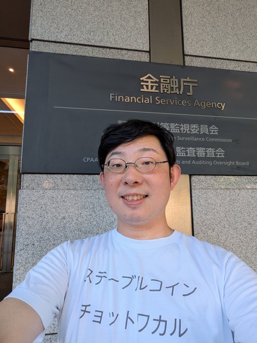 A man wearing glasses and a white t-shirt with Japanese text ("ステーブルコイン チョットワカル" - "I sort of understand stablecoins") takes a selfie in front of a sign for the Financial Services Agency (金融庁), which also references the Securities and Exchange Surveillance Commission and CPAAO. The image visually sets the scene for his upcoming visit to discuss stablecoin regulations and cryptocurrency-related matters with Japan's financial regulator.
