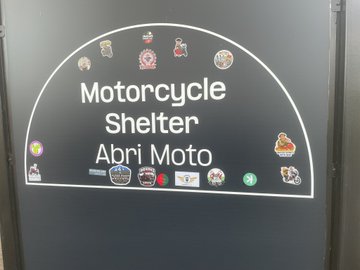 A black sign with white text reading 'Motorcycle Shelter / Abri Moto' decorated with various colorful stickers, indicating a parking area for motorcycles.