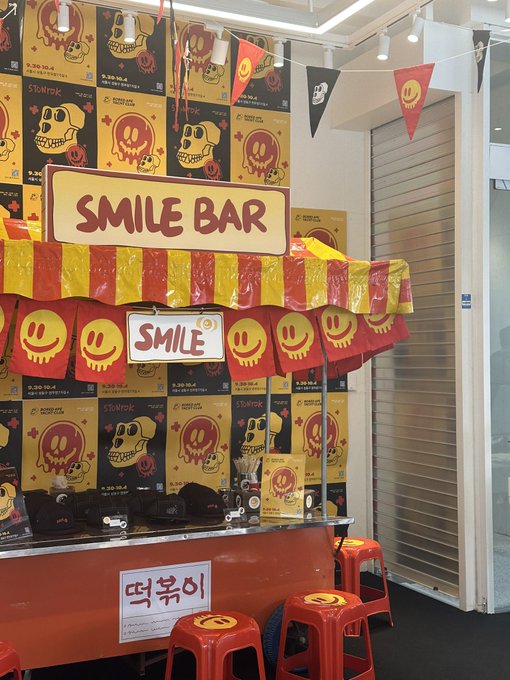 An indoor photo booth or bar stand called 'SMILE BAR' is decorated with red and yellow skull designs, smiley face garlands, and a sign with Korean text '딱볶어'.