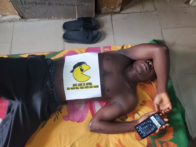 A shirtless man lies on a colorful floral mat, holding a smartphone displaying the $ARCMAN game, with a sign on his chest reading "ARC-ADE IS OPEN. ARC-MAN WILL TAKE OVER ARC-CHAIN," visually parodying the iconic Pac-Man arcade game while promoting the $ARCMAN cryptocurrency project.