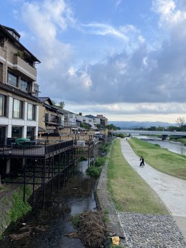 A scenic view of a riverside town with traditional Japanese-style buildings on stilts along a narrow waterway, with a wide gravel riverbed and a cloudy sky.