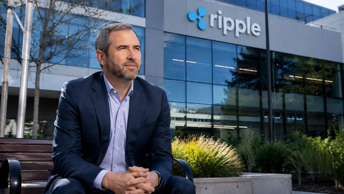 The image features a man sitting on a bench outside of Ripple headquarters. The man appears to be dressed in business attire, possibly a suit or a blue shirt with a tie. In front of him, there's a large building that houses Ripple offices. The text "Ripple has now deployed over $550M into the XRPL ecosystem" is visible on the image, indicating that Ripple has invested a significant amount of money in the cryptocurrency space. Additionally, the post mentions that Ripple will be deploying over 2026 to become its biggest expansion phase yet.