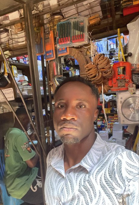A selfie of a man in a patterned white shirt standing inside a hardware store, with tools, ropes, and various supplies visible on shelves behind him.