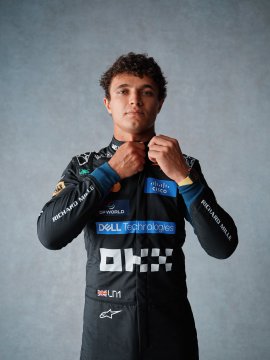 A McLaren F1 racing driver adjusts his black and blue team uniform, which prominently displays "Dell Technologies," "OKX," and "Hilton" sponsor logos alongside the iconic McLaren checkered flag emblem. The portrait captures the signature look of the team's official apparel line, fittingly described as "an iconic fit for an iconic World Champion."