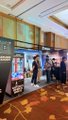 A blurry, low-resolution image showing an indoor event space or convention with several people standing around a table or counter.