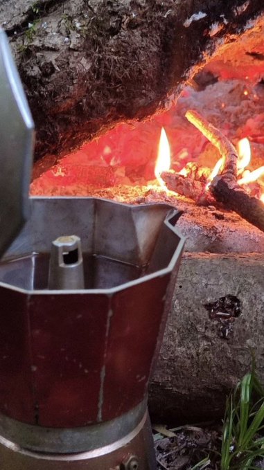 A traditional octagonal moka pot coffee maker sits near an open fire with burning logs and glowing embers.