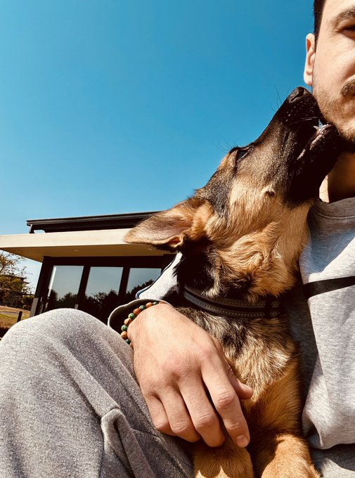 A man cuddles a German Shepherd dog outdoors under a clear blue sky.