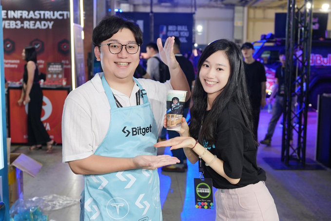 A smiling man wearing a light blue "Bitget" branded apron waves at the camera while posing with a smiling woman in a black top and pink skirt at the Bitget booth, where she holds up a promotional item. The accompanying post celebrates "FutureMode Day 2" and invites attendees to visit the Bitget booth, which directly reflects the lively booth atmosphere and branded apparel visible in the image.
