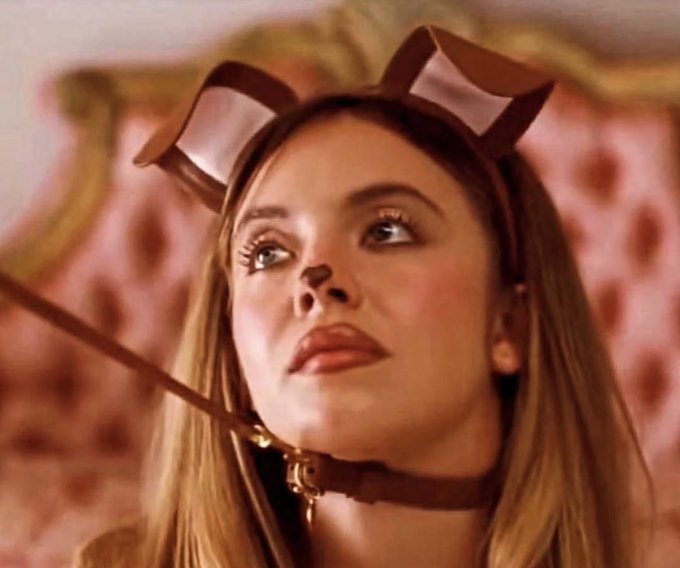 A close-up portrait of a young woman with long blonde hair wearing brown bunny ears and a matching choker, looking upwards against a pink, tufted background.