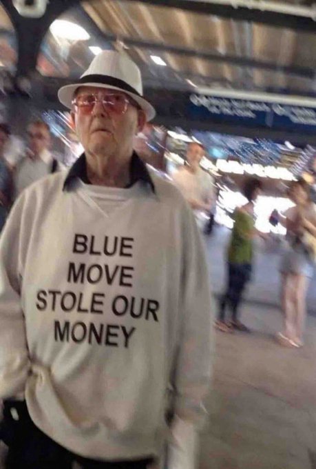 An older man wearing glasses and a white hat stands outdoors, wearing a sweatshirt with the text "BLUE MOVE STOLE OUR MONEY".