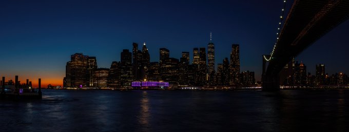 A nighttime cityscape showing the illuminated skyline across the water at dusk, with the lights of an iconic suspension bridge visible on the right side.