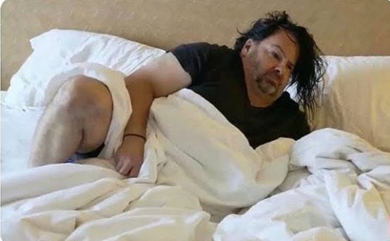 A disheveled man lies in bed with rumpled white sheets, appearing startled or bleary-eyed as if just woken up. The image humorously captures the "waking up to Bitcoin milestones" feeling referenced in the post, with his surprised expression perfectly mirroring the recurring excitement of seeing $64k Bitcoin for the 124th time.