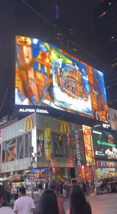 A nighttime photograph of a large LED billboard above a Popeyes and McDonald's restaurant in a busy urban area, displaying a colorful animated advertisement credited to 'ALPER ÖZDIL'.