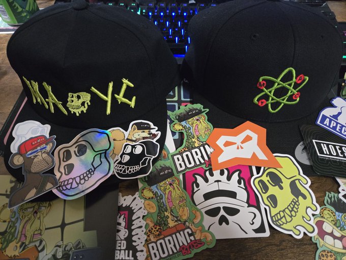 In the image, there are two baseball caps displayed side by side on a table. The cap on the left has a skull design with neon green letters spelling "Kaiju", while the cap on the right features an anime character with a green head and orange body. Both caps have various stickers attached to them, including one that reads "born" and another that says "mutant". These stickers add a personal touch to the caps, making them unique and interesting. The image also includes text elements such as "mutant merch Monday is always one of the greatest feelings especially from @KaijuTheApe & @apewhale", which suggests that these caps might be related to an event or promotion involving mutant-themed merchandise.