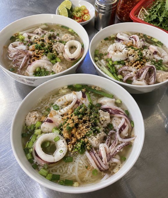 Three bowls of Vietnamese-style squid (mực) noodle soup topped with green onions, fried garlic, and pepper, served with lime wedges and chili peppers on the side.