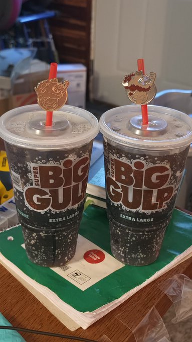 Two Super Big Gulp extra-large cups with decorative animal-shaped straw toppers sit on a green napkin on a wooden table.