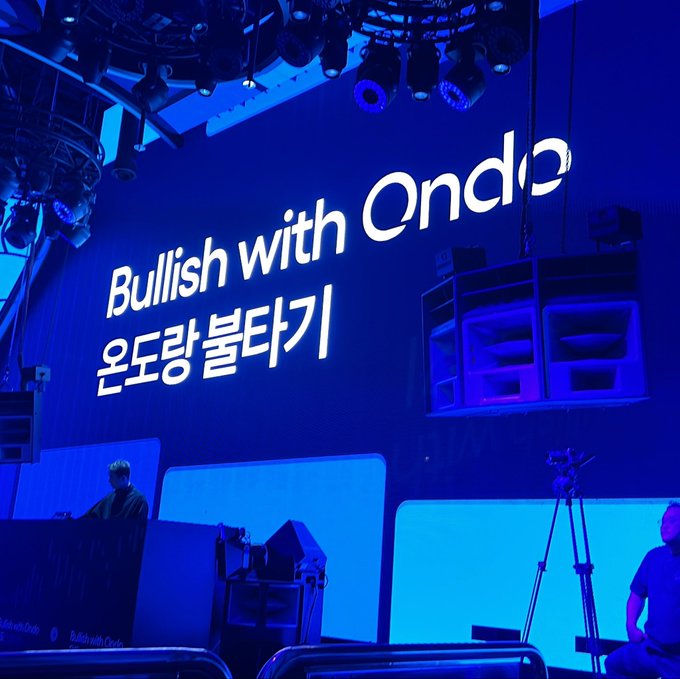 A large blue screen at an event displays the text 'Bullish with Ondo' and Korean text '온도랑 불타기', with stage lighting visible above.