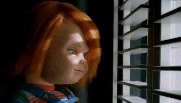 Chucky, the red-haired doll character, smiling while looking through window blinds.