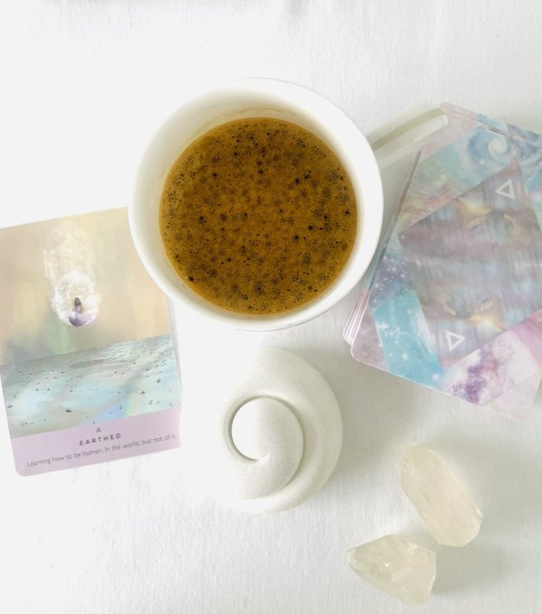 A white mug of frothy coffee or tea sits on a saucer next to an oracle card labeled 'EARTHED' with learning to be human text, alongside galaxy-themed cards and clear quartz crystals.
