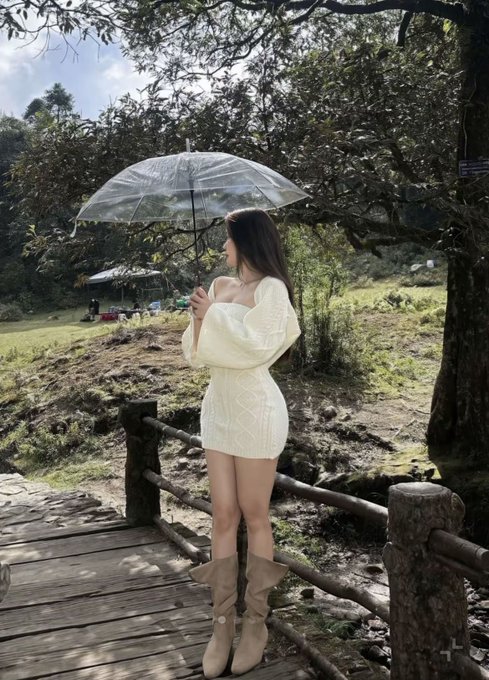 A woman in a white off-the-shoulder knit dress and tan boots standing on a wooden bridge holding a clear umbrella, surrounded by lush green trees and foliage.