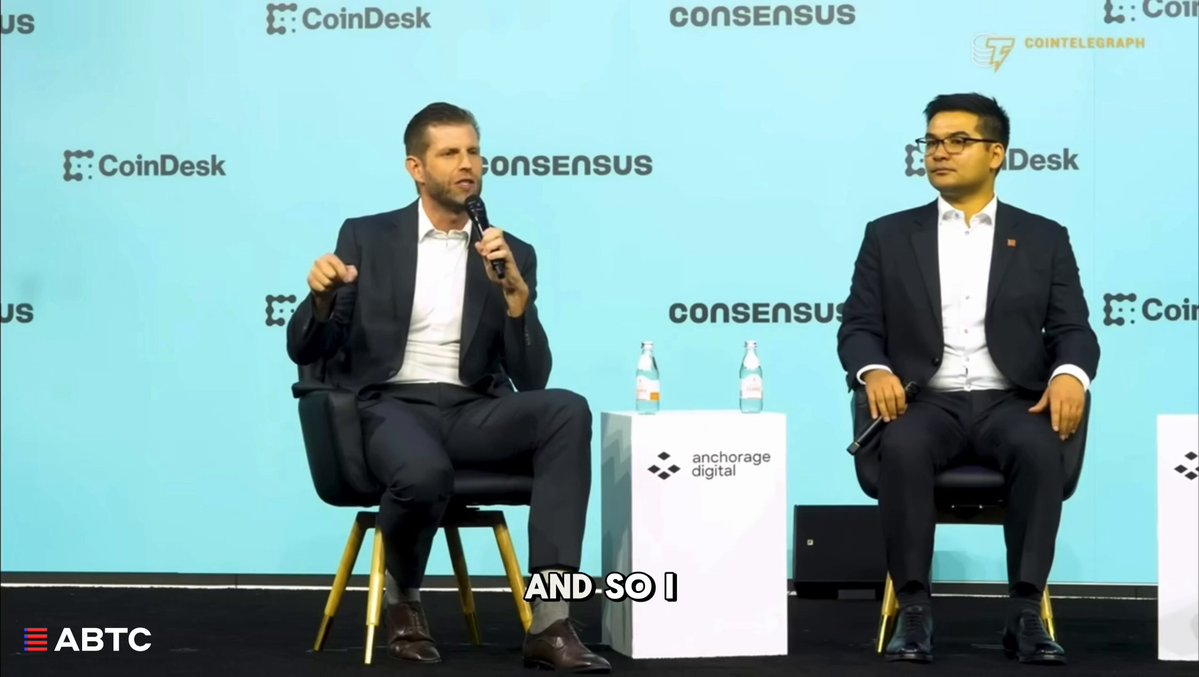 The image shows two men sitting behind a table, one wearing a suit and the other dressed more casually. They are engaged in conversation or presentation, possibly discussing Bitcoin mining. The background features a blue wall with white text that reads "Consensus".