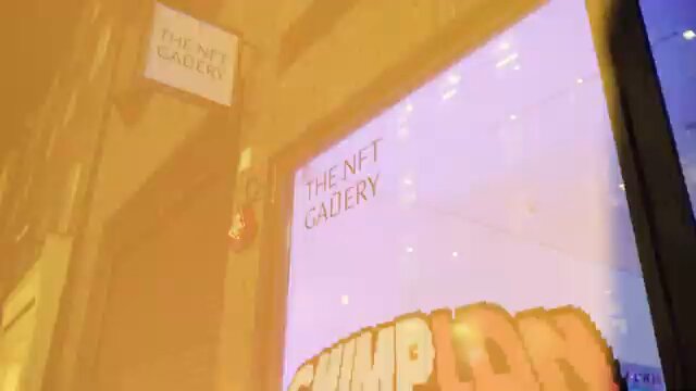 The image shows a store window with a neon sign for "The NFT Gallery". The neon sign displays the words "The NFT Gallery" written in white letters against an orange background. This eye-catching display helps attract customers' attention, making them curious about what is inside the store and encouraging them to explore further.