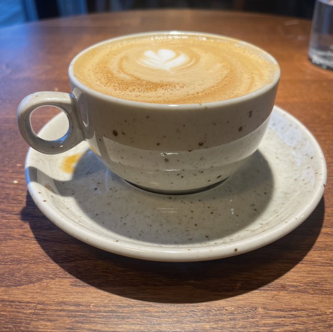 A speckled ceramic cup of cappuccino with heart-shaped latte art sits on a matching saucer on a wooden table.