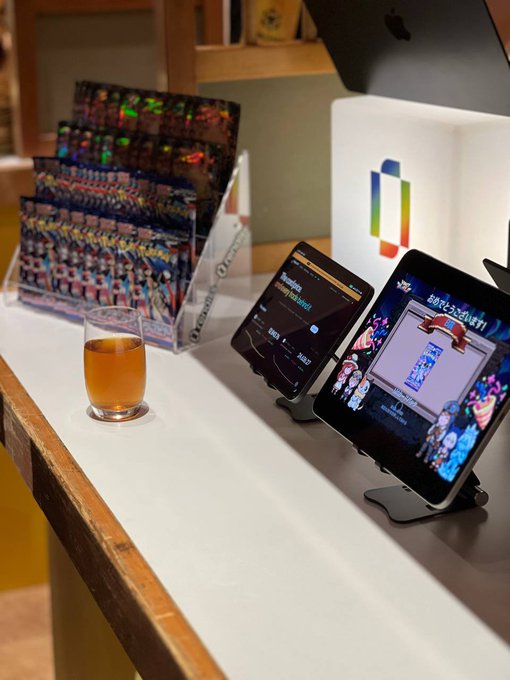 A counter displaying a small glass of tea, a brochure rack with what appears to be 'Orientalradio' flyers, and two tablets on stands showing Japanese content including a Pokémon game, with a colorful square logo on the wall behind.