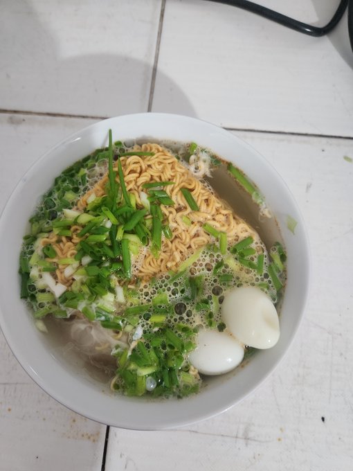 The image features a bowl of food, possibly ramen noodles with an egg, sitting on top of a table. The bowl contains green onions as well, adding color and flavor to the dish. The photo appears to be taken from a low angle, making the bowl look more prominent in the frame. The post that accompanies this image is titled "Ăn mì tôm đi quảng các dự án triệu đân giờ đây, người dùng có thể chạy các tác vụ tối ưu hóa nhỏ trực tiếp". The image seems to be promoting a dish with noodles and an egg, possibly ramen noodles with an egg.