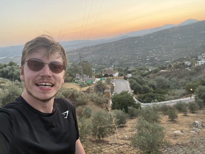 A young man wearing sunglasses and a black Gymshark t-shirt takes a selfie outdoors with a sunset over hills and a village in the background.