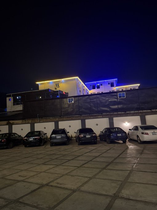 A nighttime view of a building with yellow and blue neon lights, with several cars parked in front.