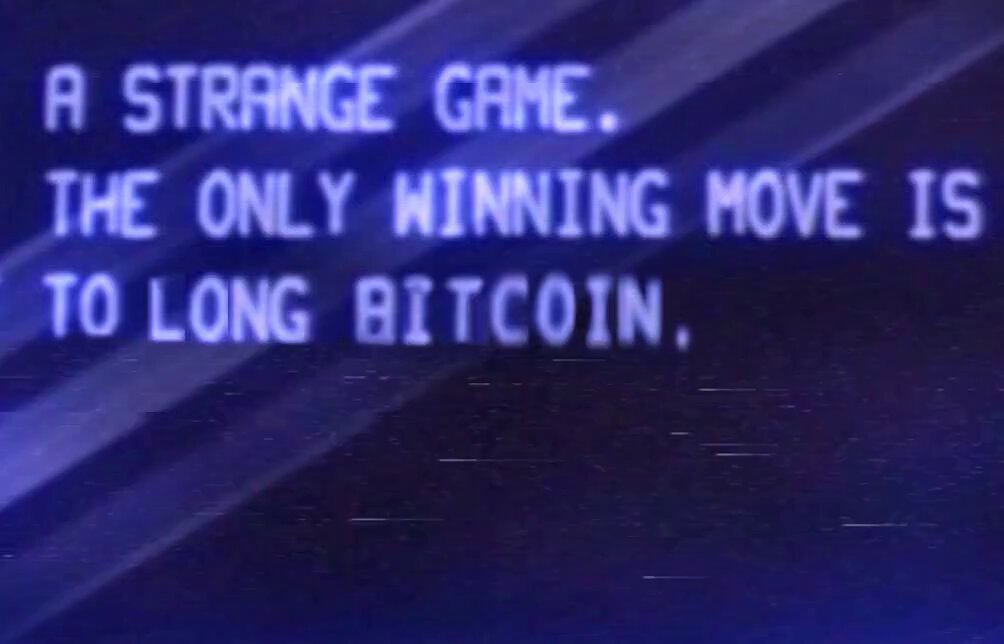 The image features a blue sign with white text that reads "A Strange Game. The only winning move is to long bit coin." This message seems to be related to a discussion about Bitcoin, as indicated by the reference to "TezArt" in one sentence and "DigitalArt" in another. The sign appears to be posted on social media or an online platform where people share information about various topics.