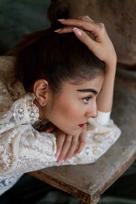 Profile portrait of a woman with her hair in a top bun, resting her chin on her hand while wearing a delicate white lace top.