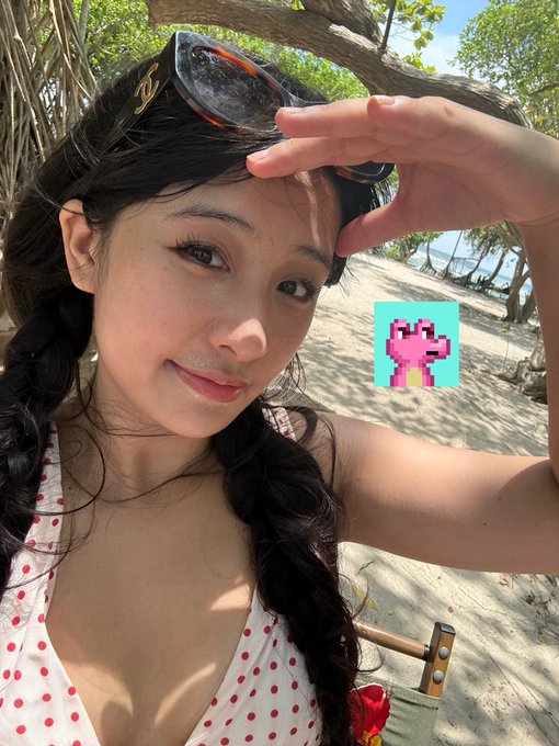 A woman with a long braid poses for a beach selfie in a polka-dot swimsuit, shielding her eyes from the sun, with a pixel-art pink axolotl sticker added to the scene—playfully fitting the "GMGN GO & Bestin" crypto theme.