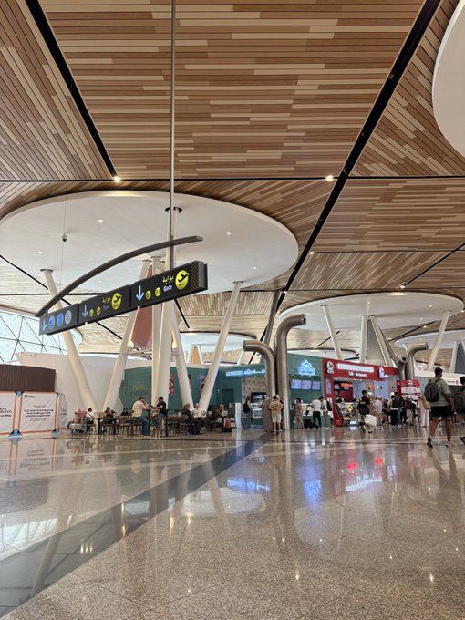 An interior view of a modern airport terminal features a curved wooden ceiling, directional signage hanging from the ceiling, and passengers walking or seated in the background.