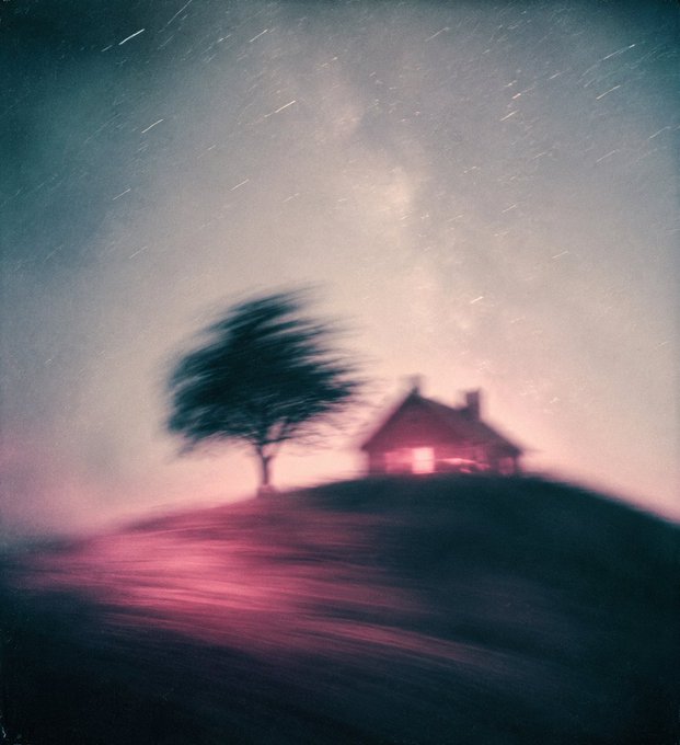 An artistic long-exposure photograph showing a small lit house and a tree on a hill under a starry night sky with the Milky Way visible.