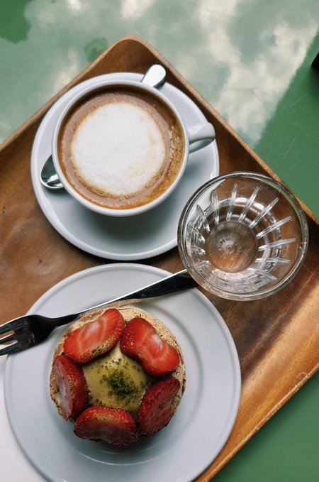 An overhead view of a wooden tray holding a cappuccino with foam art, a glass of water, and a small dessert topped with fresh strawberries and pistachio.
