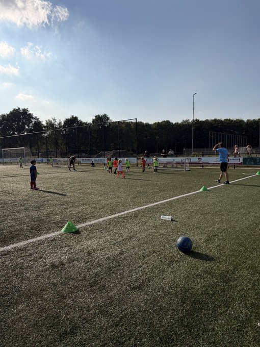 Children play soccer on a turf field during golden hour, with a blue ball resting near the foreground cone and players gathered around the goal in the distance, capturing the essence of a perfect evening away from the markets.