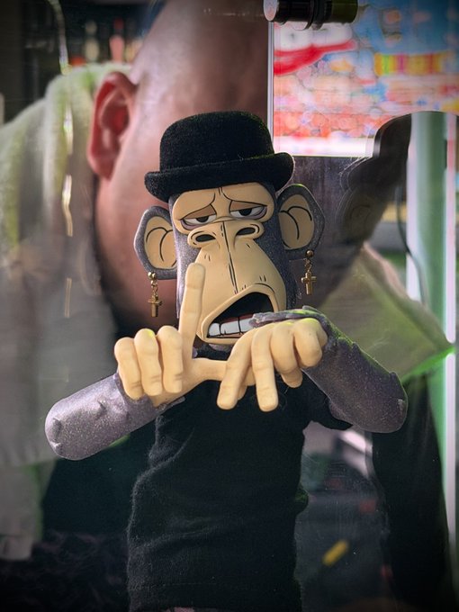 A plush monkey figure wearing a black bowler hat and cross earrings is displayed behind glass, posed with its hands near its mouth as if biting its nails, set against the reflection of a person and colorful artwork in the background, fitting the post's celebration of sharing their art with "ape friends" in LA.