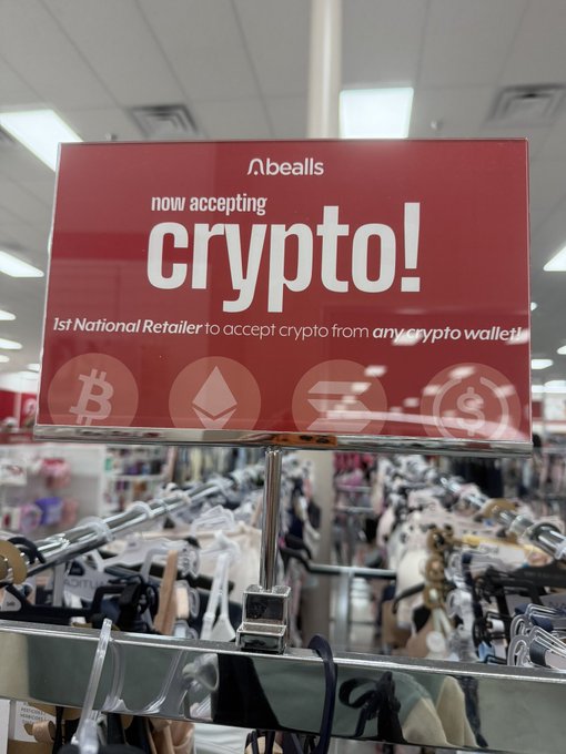 A red sign inside a Bealls department store reads "now accepting crypto! 1st National Retailer to accept crypto from any crypto wallet!" featuring Bitcoin, Ethereum, Solana, and USDC logos, confirming the poster's excitement that crypto payments have arrived at their local retailer.