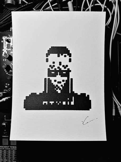 A pixel art portrait of a bearded man is printed on white paper resting on audio equipment and cables, with a signature in the bottom right corner.