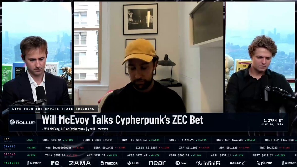 The image shows three men sitting at a table with a TV screen behind them displaying a news segment about Bitcoin and cryptocurrency. The first man, wearing a black suit, sits next to another man who is wearing a yellow hat. The third man, also in a black suit, sits further back from the other two. They are all focused on the television screen as they watch the news.